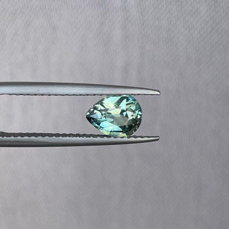 May include: A pear-shaped, faceted gemstone with shades of teal and green, held by a pair of silver tweezers. The gem has a brilliant cut, reflecting light. The background is a plain, light gray.