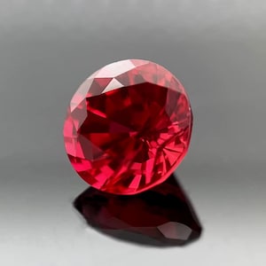 May include: A faceted, oval-shaped, ruby gemstone. The gem is a deep, saturated red color, with multiple facets that reflect light. The gem is set against a dark background, with its reflection visible below.