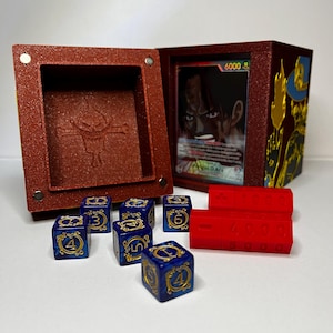 May include: A red textured box with a magnetic closure, open to reveal a card and a compartment. Six blue dice with gold numbers and symbols are next to the box, along with two red counters. The card features a character illustration.