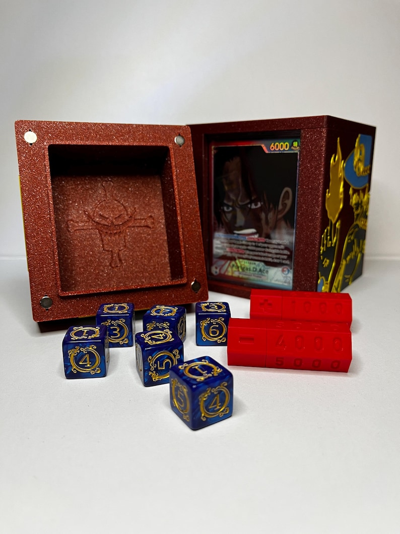 May include: A red textured box with a magnetic closure, open to reveal a card and a compartment. Six blue dice with gold numbers and symbols are next to the box, along with two red counters. The card features a character illustration.
