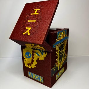 Può includere: Una scatola marrone con coperchio incernierato, con caratteri giapponesi gialli e un'emblema dorato. La scatola ha una finitura strutturata e un ritratto di un personaggio con cappello blu e dettagli dorati. La parola "AXCE" è stampata.