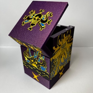 Puede incluir: Una caja de almacenamiento cuadrada de color morado con tapa abatible. El exterior presenta diseños gráficos en relieve en dorado y azul, incluyendo un emblema de calavera y tibias cruzadas en la tapa y motivos de criaturas con cuernos.
