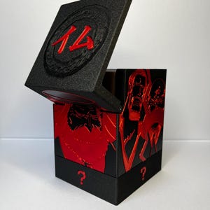 Può includere: Scatola nera con design rosso e coperchio leggermente aperto. Il coperchio presenta un carattere giapponese rosso in un cerchio nero. I lati della scatola presentano disegni in rilievo rossi e punti interrogativi. La scatola è su una superficie bianca.