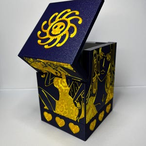 Può includere: Una scatola blu navy con coperchio incernierato, con disegni in rilievo color oro. Il coperchio mostra un emblema a forma di sole, mentre i lati presentano figure e dettagli a forma di cuore. La scatola è aperta.