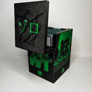 Könnte beinhalten: Schwarze Kartenaufbewahrungsbox mit grünen Akzenten. Die Box hat einen Klappdeckel mit grünen japanischen Schriftzeichen. Die Seiten der Box weisen grüne Illustrationen und dekorative Elemente auf. Für Sammelkarten.