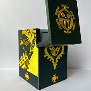 Può includere: Una scatola verde scuro con coperchio incernierato, con disegni grafici color oro. La scatola presenta un'illustrazione gialla e nera sul lato. Il coperchio è aperto, rivelando l'interno. La scatola ha un emblema dorato sul coperchio.