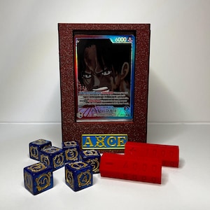 May include: A framed trading card featuring a character with dark hair and a determined expression. The frame is a deep red color, and the card is displayed with blue dice and red building blocks. The text "AXCE" is visible.