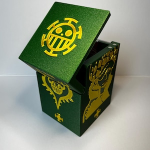 May include: A dark green, glittery, rectangular storage box with a hinged lid. The lid features a yellow circular emblem with a skull-like design. The front and side panels display yellow graphic illustrations of characters and symbols.