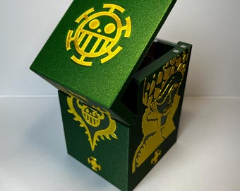 May include: A dark green, glittery, rectangular storage box with a hinged lid. The lid features a yellow circular emblem with a skull-like design. The front and side panels display yellow graphic illustrations of characters and symbols.