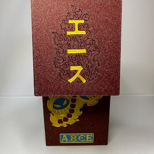 May include: A red, textured box with yellow Japanese characters and a flame design. The bottom of the box features a gold emblem with blue accents and the word "ACE" in a blue box.