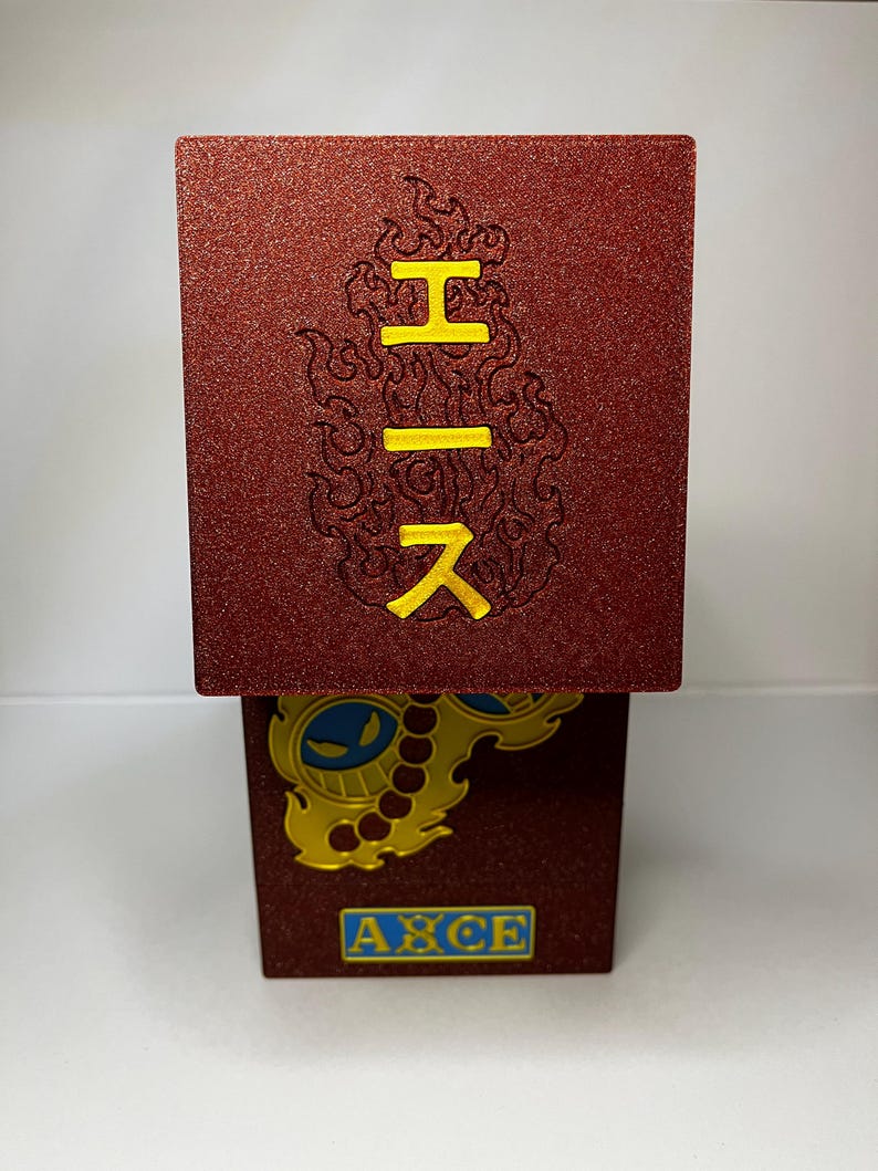 May include: A red, textured box with yellow Japanese characters and a flame design. The bottom of the box features a gold emblem with blue accents and the word "ACE" in a blue box.