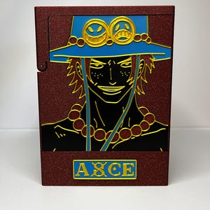 May include: A maroon cigarette case featuring a detailed illustration of a character with a blue hat and gold accents. The character's face is black with gold outlines, and the word "ACE" is displayed in a gold rectangle.