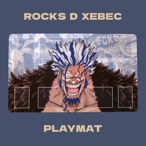 May include: A rectangular playmat featuring a central illustration of the character Rocks D Xebec with spiky white and blue hair, a dark feathered collar, and a wide grin. The background displays faint manga-style panels. Text reads ROCKS D XEBEC PLAYMAT.