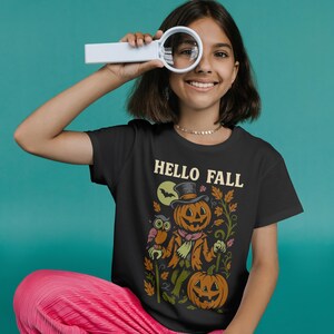 Może przedstawiać: Czarny t-shirt z motywem Halloween. Projekt przedstawia postać dyni w kapeluszu i szaliku, z napisem "HELLO FALL". Koszulka jest połączona z różowymi spodniami.