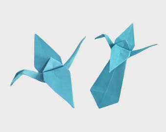May include: Two origami cranes in flight, crafted from light blue paper. The birds are depicted with wings spread, showcasing the intricate folds and shapes of the paper art. The background is a solid, neutral color.