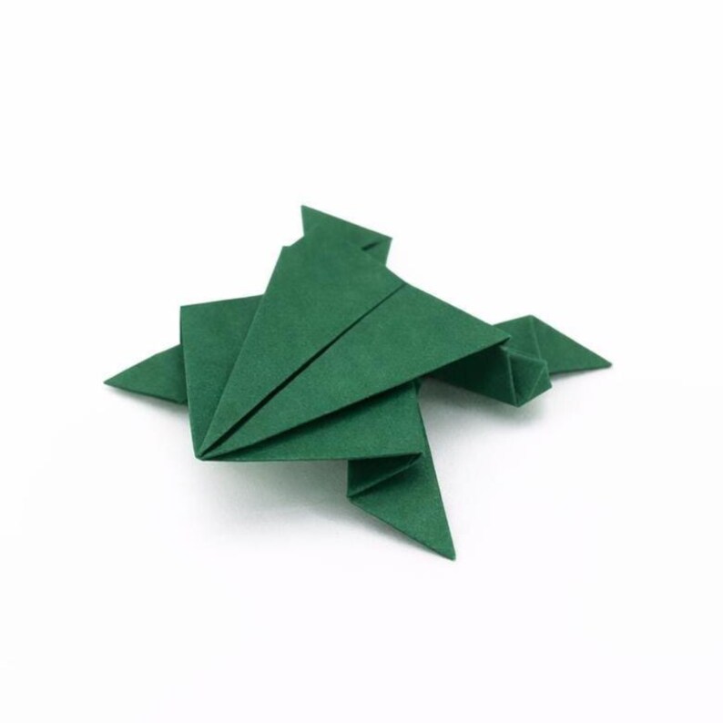 May include: A green origami frog, crafted from folded paper, sits on a white background. The frog's body is composed of geometric shapes, with triangular legs and a square body. The paper has a matte finish.