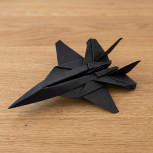 May include: A black origami paper airplane folded into the shape of a fighter jet. The model features a pointed nose, swept-back wings, and a distinct tail section with vertical stabilizers, all constructed from a single sheet of matte black paper.