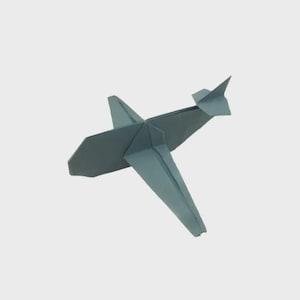 May include: A folded paper airplane in shades of blue-gray. The origami plane has wings, a tail, and a pointed nose, all created from folded paper. The plane is angled slightly, suggesting flight, against a plain background.