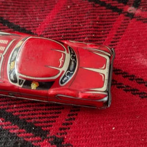 Peut inclure: Une voiture jouet rouge vintage avec des accents argentés et blancs. La voiture classique présente un design détaillé, comprenant un toit, des fenêtres et des éléments décoratifs. La voiture jouet est sur une surface à carreaux rouge et noir.