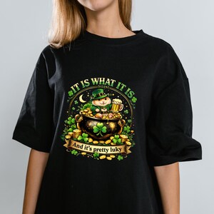 Può includere: T-shirt nero con grafica per la festa di San Patrizio. Il design include un folletto in un pentolone d'oro, un boccale di birra, trifogli e le scritte "It is what it is" e "And it's pretty luky".