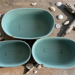 Può includere: Tre cesti ovali in corda turchese di diverse dimensioni. I cesti presentano un design a cucitura a spirale e piccoli dettagli in pelle marrone. Conchiglie e legno galleggiante sono disposti su una superficie di legno.