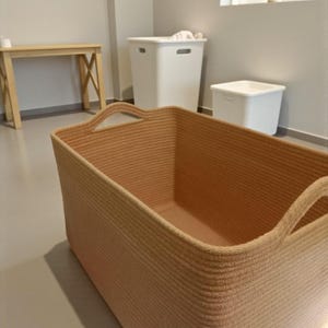 Puede incluir: Una cesta de almacenamiento rectangular grande, tejida en color beige claro, con asas integradas. La cesta está colocada sobre un suelo gris claro, con una mesa de madera y dos contenedores de almacenamiento blancos en el fondo. La habitación tiene un diseño minimalista.