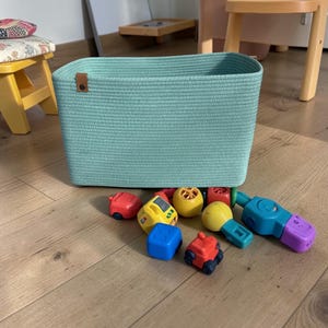 Könnte beinhalten: Ein rechteckiger, helltürkisfarbener Aufbewahrungskorb mit einem braunen Lederanhänger. Verschiedene bunte Spielzeugautos und -blöcke liegen auf dem Holzboden vor dem Korb. Ein gelber Hocker und ein kleiner Tisch sind im Hintergrund.