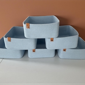 May include: Six light blue woven storage baskets with square shapes are arranged on a white surface. Each basket has a brown leather-like tag on the front. The background is a solid brown color.