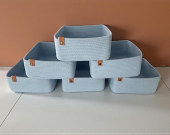May include: Six light blue woven storage baskets with square shapes are arranged on a white surface. Each basket has a brown leather-like tag on the front. The background is a solid brown color.