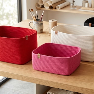 May include: Three rectangular storage baskets made of coiled rope, featuring a red, a pink, and an off-white basket. Each basket has a small tan leather-like tab attached to the rim with a metal rivet, providing a structured and open design.