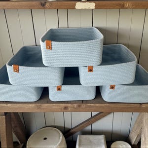 Peut inclure: Un ensemble de six paniers de rangement rectangulaires bleu clair fabriqués en corde torsadée. Chaque panier présente une languette en similicuir marron fixée sur le bord avant avec un rivet métallique, offrant un aspect uniforme à l'ensemble.