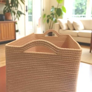 Puede incluir: Una cesta rectangular tejida, de color marrón claro, con dos asas. La cesta está hecha de hebras horizontales, muy tejidas, creando una superficie texturizada. El color neutro y el diseño sencillo sugieren un artículo de almacenamiento o decoración.