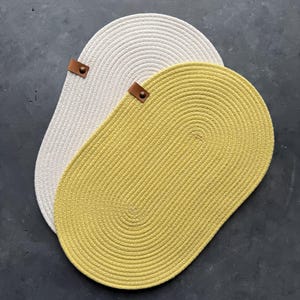 Op de afbeelding: Twee ovale placemats gemaakt van gewikkeld touw, één in een lichte crèmekleur en de andere in een mosterdgele tint. Elke placemat is voorzien van een klein bruin leren label dat met een metalen klinknagel aan de rand is bevestigd.
