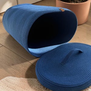 Op de afbeelding: Een diepblauwe ovale opbergmand gemaakt van gedraaid touw met een bijpassend deksel voorzien van een centrale handgreep. De mand heeft een klein bruin lederachtig lipje aan de bovenrand. De structuur is flexibel en hol met een donkere binnenkant.