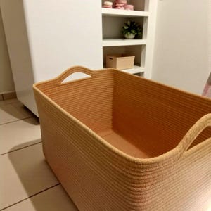 Peut inclure: Un panier de rangement rectangulaire fabriqué en corde beige enroulée, doté de poignées intégrées sur deux côtés. Le panier présente un intérieur profond et ouvert ainsi qu'une surface texturée formée par des rangées horizontales continues.