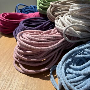 Puede incluir: Una colección de cordones de algodón enrollados en varios colores, incluyendo azul claro, rosa suave, blanco, beige, morado, magenta, azul oscuro y verde claro. Cada cordón presenta una textura trenzada y está enrollado en un haz.