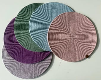 May include: A set of five circular placemats made from coiled rope in various colors including light purple, deep purple, sage green, light blue, and dusty pink. Each placemat features a consistent spiral texture and a small brown tag.