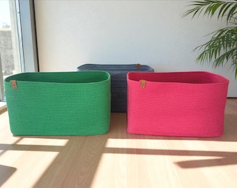 May include: Three rectangular woven storage baskets in green, gray, and pink. Each basket has a rectangular shape and a small leather tag. The baskets are on a wooden floor, with a window and a plant in the background.