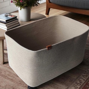 May include: A beige rectangular storage basket made of woven material. The basket has a brown leather accent and sits on small black wheels. A round wooden table with books and a vase of flowers is in the background.