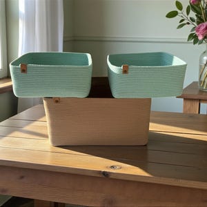 Puede incluir: Tres cestas de almacenamiento rectangulares hechas de cuerda enrollada. Dos cestas más pequeñas son de color verde menta claro y una cesta más grande es de color tostado. Cada cesta tiene una pequeña lengüeta de cuero marrón en el borde.