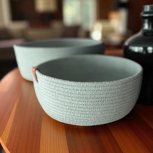 May include: Two light grey woven bowls with a textured surface. One bowl has a small brown leather accent. The bowls are on a wooden table, with a dark bottle in the background. Home decor.