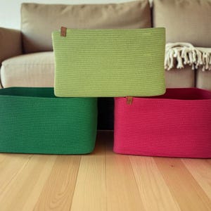 May include: Three rectangular storage baskets in shades of green and pink. The baskets are made of a woven material and have a rectangular shape. Each basket has a small brown leather tag attached. The baskets are stacked on a wooden floor.