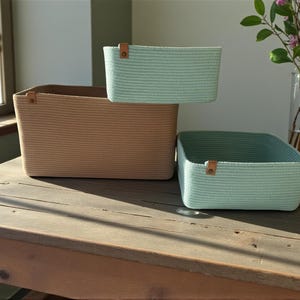 Peut inclure: Trois paniers de rangement rectangulaires en corde tressée. Un grand panier est beige, tandis que deux paniers plus petits sont vert menthe clair. Chaque panier comporte une petite languette en similicuir marron fixée sur le bord supérieur.