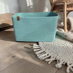 Puede incluir: Una cesta de almacenamiento rectangular hecha de cuerda enrollada de color verde azulado claro con una textura suave y uniforme. La cesta cuenta con una pequeña lengüeta de piel sintética marrón unida a la esquina superior izquierda con un remache.