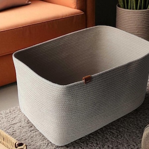 May include: A set of woven storage baskets in different sizes and shapes. The largest is a light grey rectangular basket with a small brown leather tag. Other baskets are round, and a rectangular tray is also present, all in neutral colours.