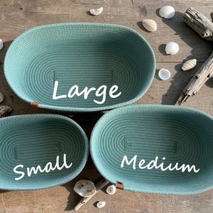 May include: Three oval-shaped sage green rope baskets in small, medium, and large sizes. Each basket features a coiled construction with a small brown leather-like tab on the rim. The text Large, Medium, and Small is printed on each.