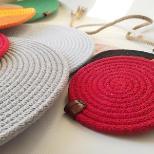 May include: A selection of circular coasters in various colours, including red, grey, orange, and green. They are made from woven rope and some have a small leather tag. These coasters are designed to protect surfaces from heat and moisture, and are approximately 10 cm in diameter.