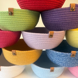 Könnte beinhalten: Eine Sammlung runder Körbe aus gewickeltem Seil in verschiedenen Farben, darunter Rot, Limettengrün, Lila, Magenta, Lavendel, Gelb, Senfgelb, Marineblau, Weiß, Hellblau und Pink. Jeder Korb hat eine kleine braune Lederlasche mit einer Metallniete.