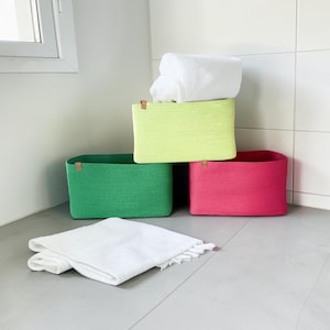 May include: A set of brightly coloured woven storage baskets in green, pink, and lime green, arranged in a bathroom. A white folded towel sits on top of a basket, with another on the floor.
