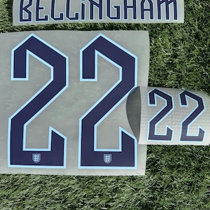 Puede incluir: Un conjunto de calcomanías para camiseta de fútbol azul marino y celeste, con el nombre "BELLINGHAM" y el número "22". Las calcomanías presentan el escudo de la selección inglesa. Sobre un fondo gris, sobre hierba verde.